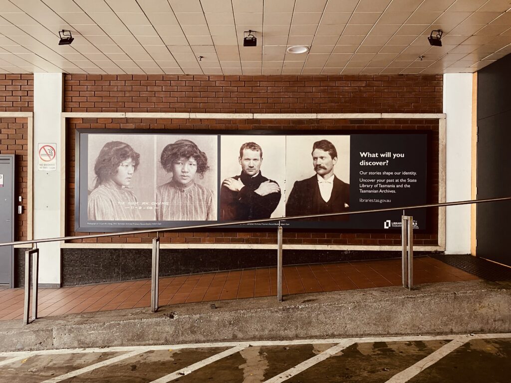 Colour photograph of a red brick wall with a large billboard on which there are old black and white portraits of an Asian woman and a white man, next to text advertising the State Library of Tasmania and the Tasmanian Archives