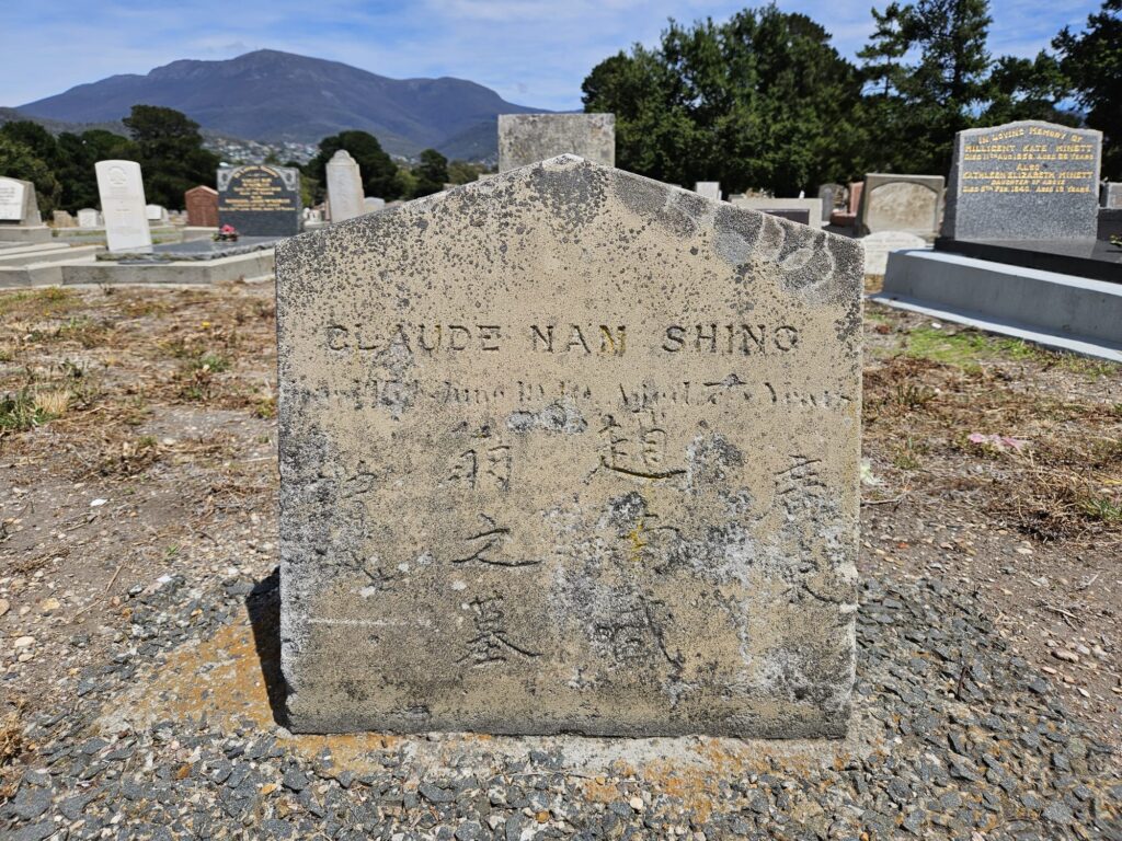 Colour photograph of a weathered stone headstone, with trees and mountains and blue sky in the background. There is English and Chinese writing on the headstone, but it is hard to read.